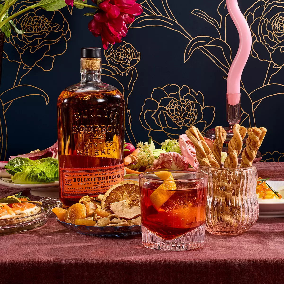 A bottle of Bulleit Bourbon whiskey next to a cocktail, breadsticks in a glass, dried fruit, salad, and other dishes on a table with a dark floral-patterned background.