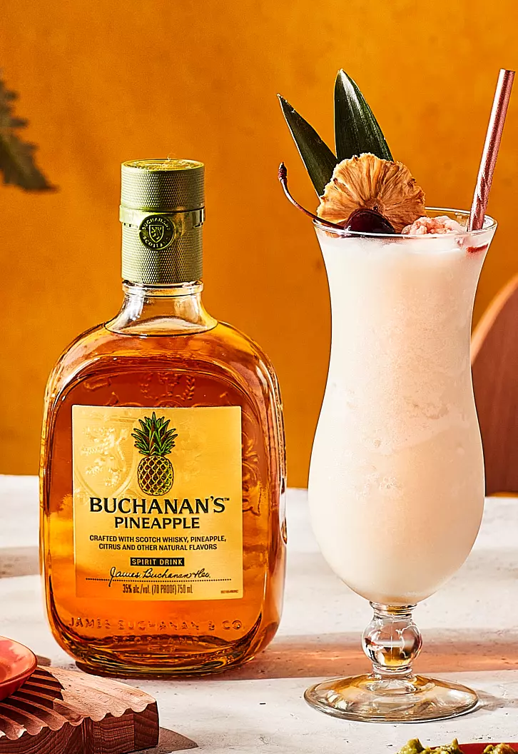 A bottle of Buchanan’s Pineapple Scotch Whisky beside a tropical cocktail garnished with pineapple, leaf, cherry, and a straw on a white surface against an orange background.