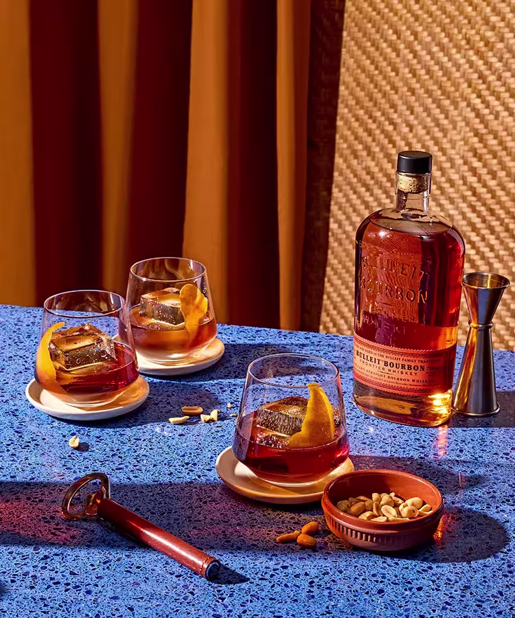 A bottle of Bulleit Bourbon, three glasses with bourbon and orange peel, a jigger, a bowl of mixed nuts, and a cigar cutter on a blue speckled table.