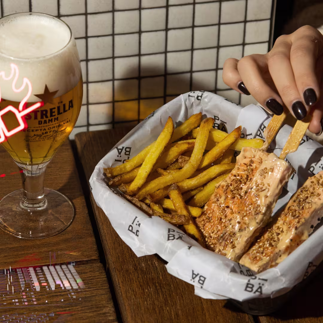 La foto muestra un primer plano de la mano de una persona sosteniendo un sandwich,sobre un recipiente de papel lleno de papas fritas. Al lado del recipiente hay un vaso de cerveza con espuma en la parte superior, etiquetado “Estrella Damm”.