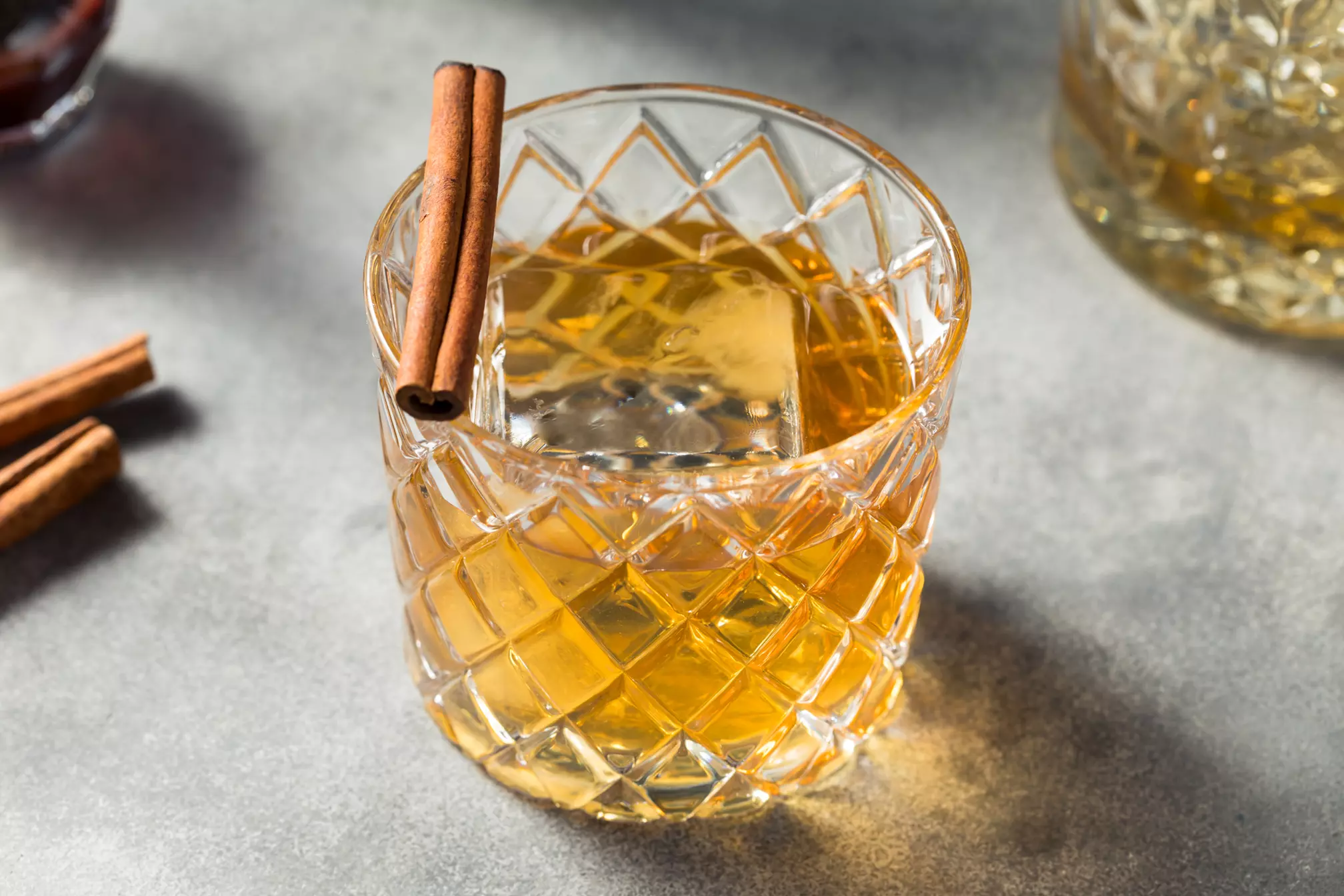 A glass of amber-colored whiskey with a large ice cube, garnished with a cinnamon stick, sits on a gray surface.