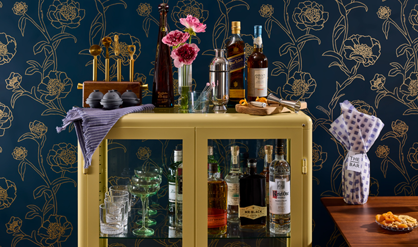 A yellow bar cart with various liquor bottles, glassware, a vase of flowers, bar tools, and a wrapped bottle sits against a dark blue floral wallpaper background.