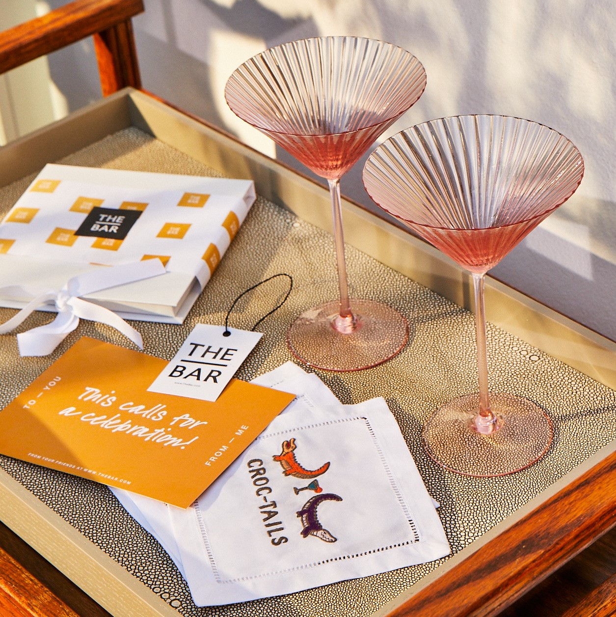 Two martini glasses with pink drinks on a tray with a gift box, a tag reading The Bar, a card stating This calls for a celebration!, and two cocktail napkins with fox designs.
