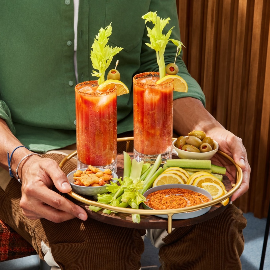 A person holds a gold tray containing two Bloody Mary cocktails and assorted garnishes such as celery stalks and green olives