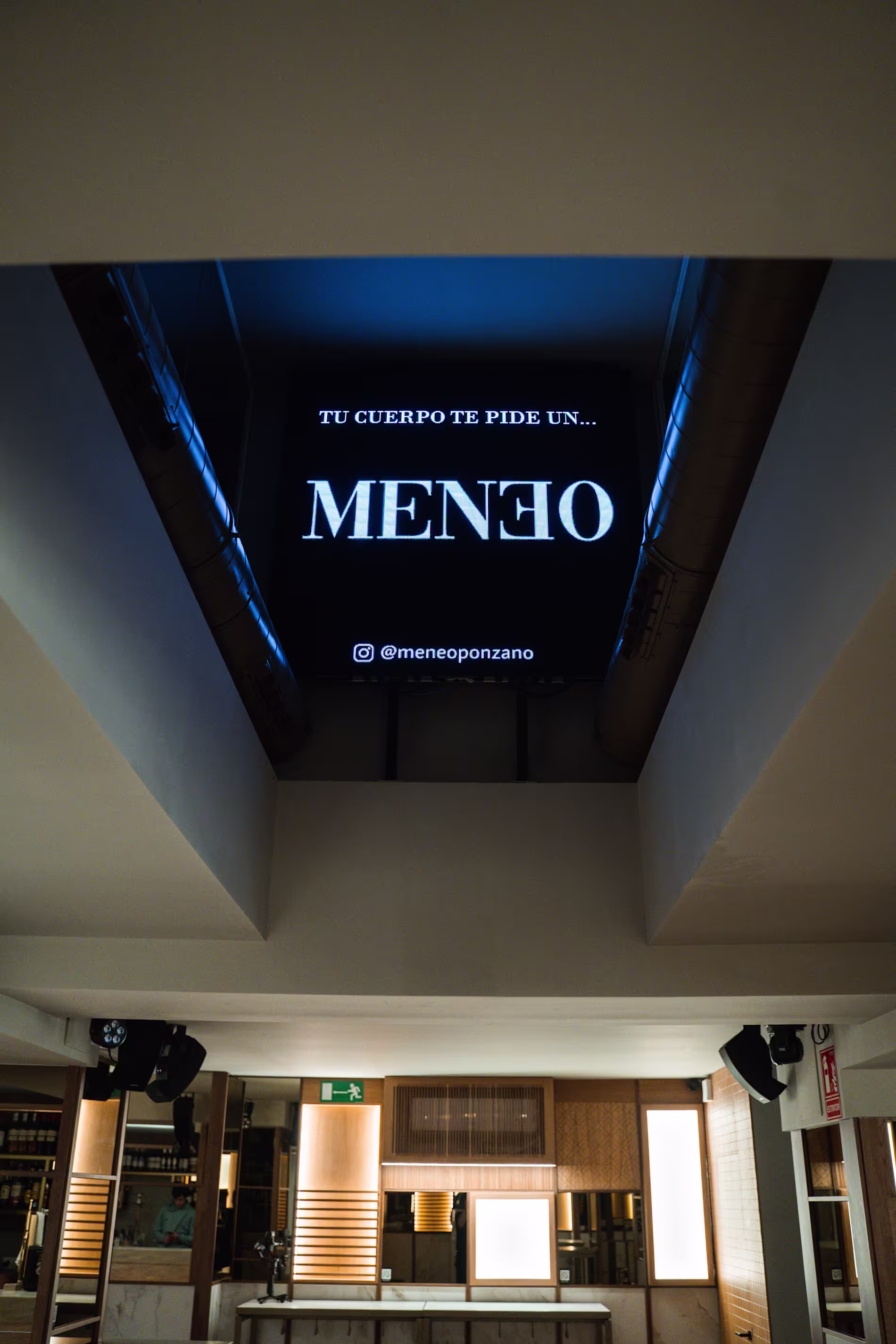 La imagen muestra el espacio interior de Meneo. se observan varias mesas iluminadas por 3 planchas de luces, la iluminiación es tenue. En el techo se observa una lamina con un letrero que MENEO.