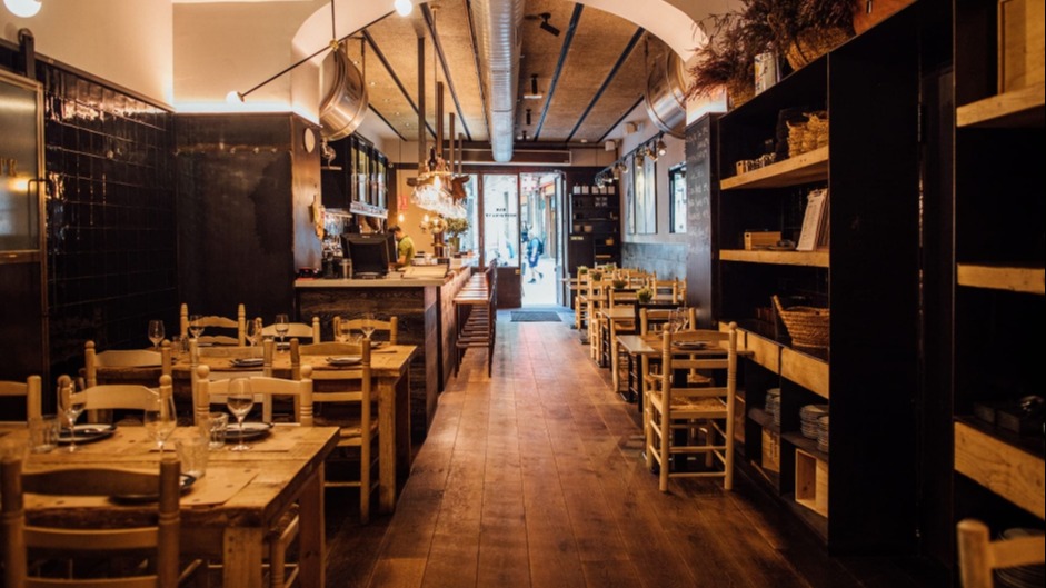 La foto muestra el interior de Quillo bar. El espacio se caracteriza por mesas y sillas de madera dispuestas ordenadamente a ambos lados de un pasillo central, que conduce a una zona iluminada al fondo que parece ser la entrada.