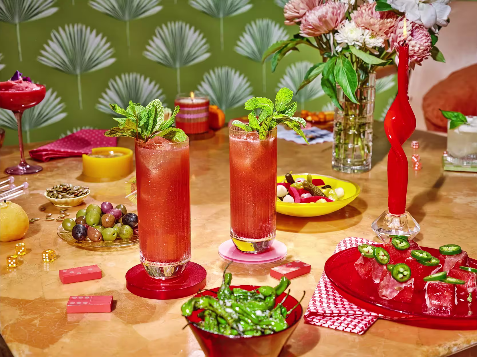 Two tall glasses of red drinks with mint garnish on a table with assorted snacks, ice cubes with jalapeños, flowers, and vibrant decor in the background.