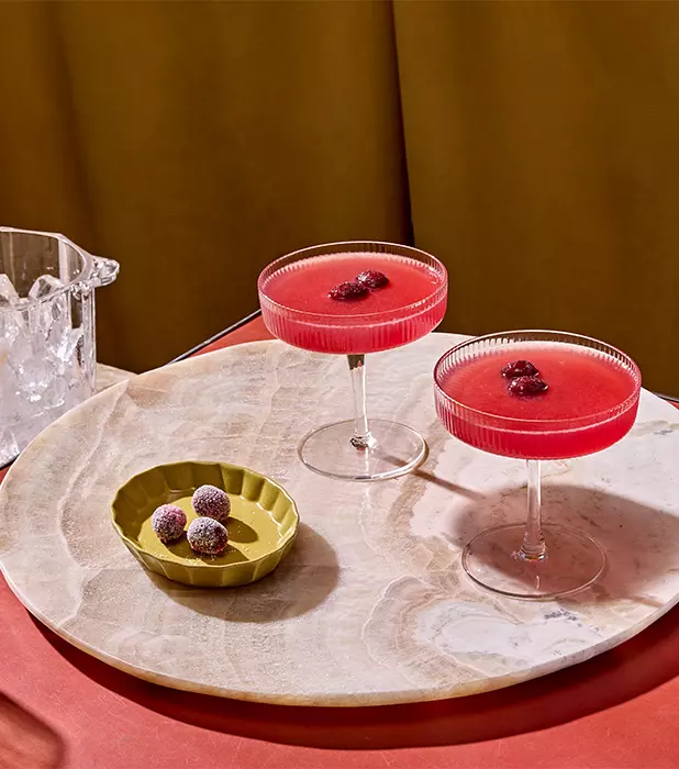 Two coupe glasses filled with a red cocktail, each garnished with berries, next to a green dish with sugared berries on a round marble tray.
