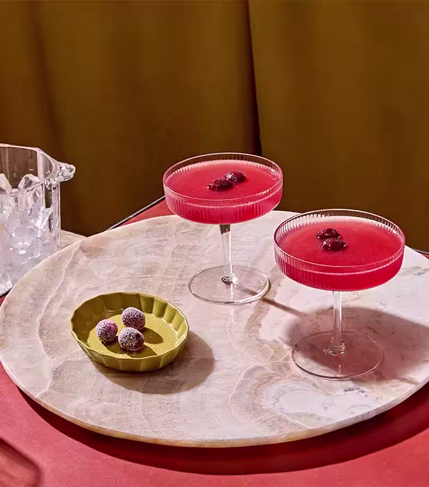 Two coupe glasses filled with a red cocktail, each garnished with berries, next to a green dish with sugared berries on a round marble tray.