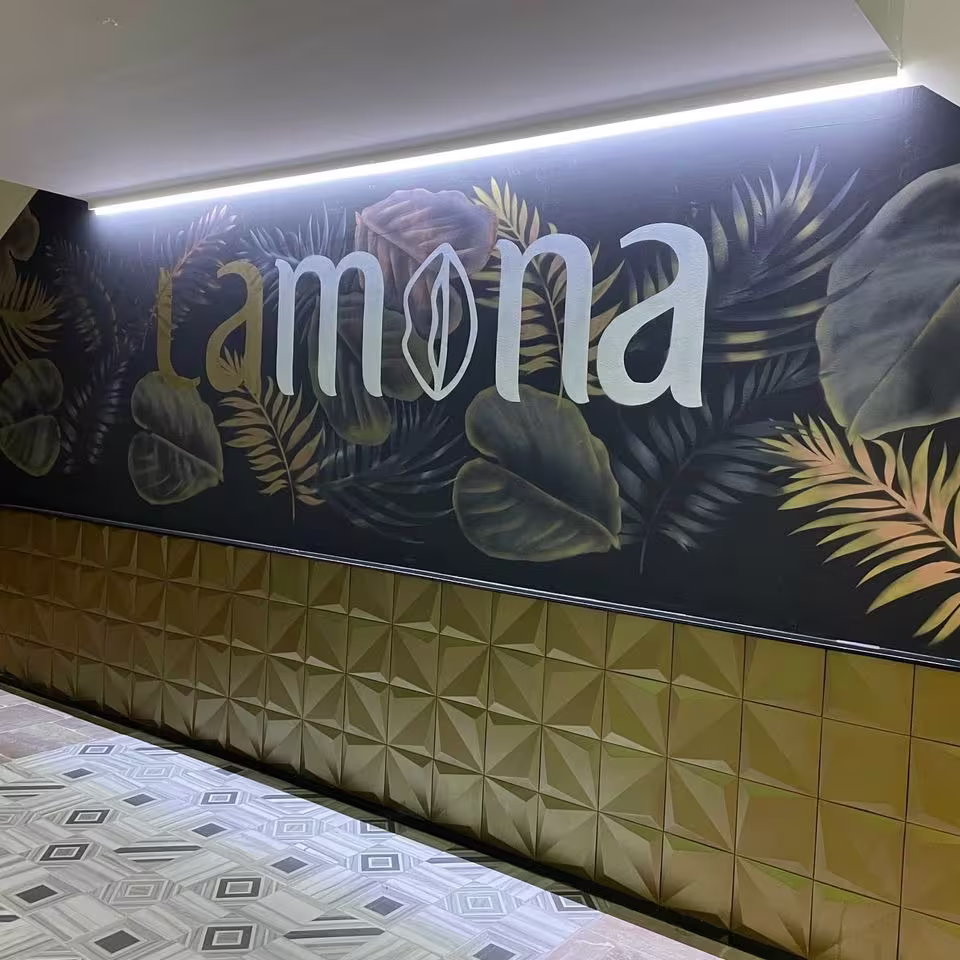 Entrada elegante con mural botánico en tonos verdes y dorados, destacando el nombre del local. Decoración moderna y cálida, con detalles en relieve dorado en las paredes, creando un ambiente sofisticado y acoged