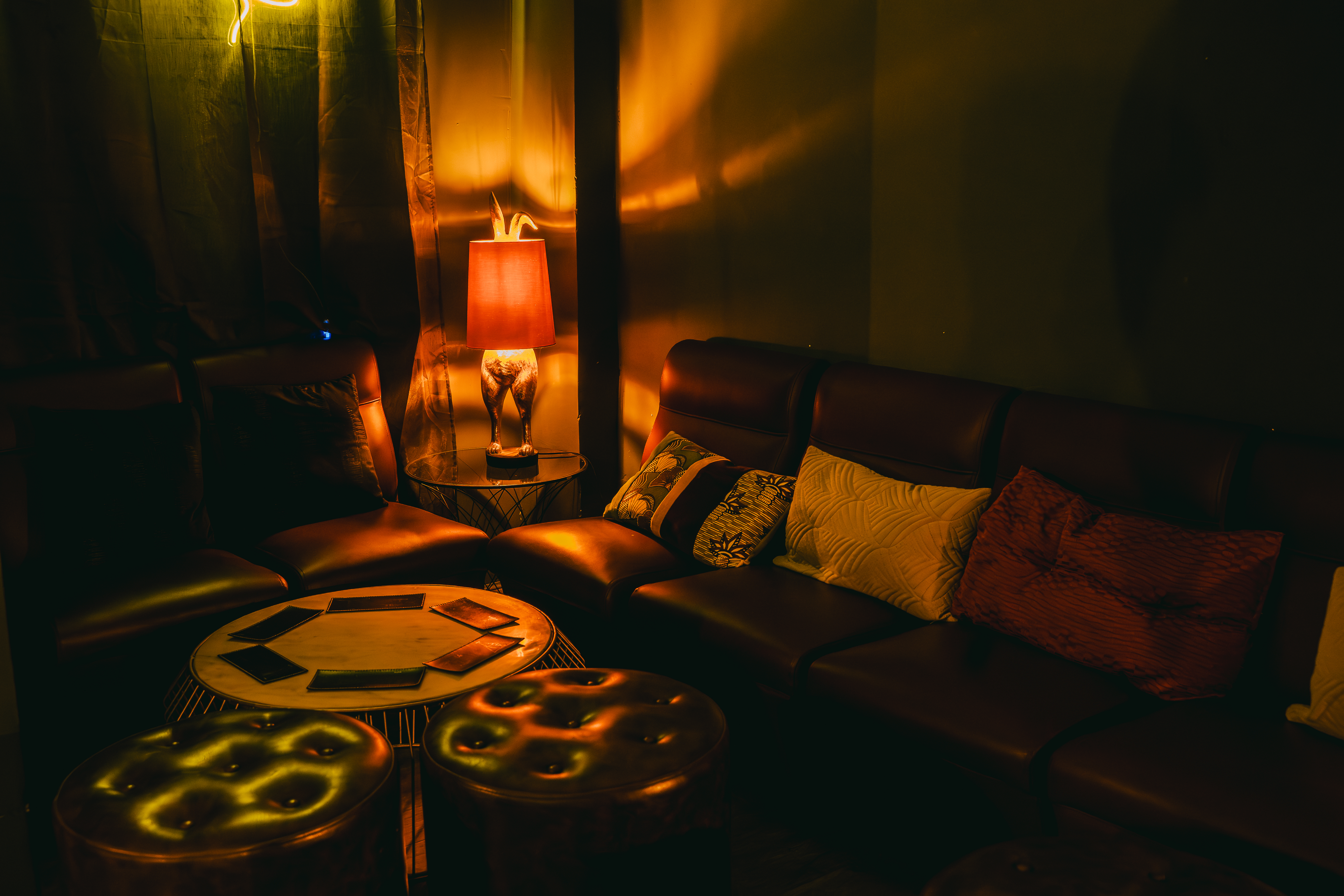 La imagen muestra una mesa del local Momus con una iluminación cálida. Hay un sofá de cuero marrón adornado con varios cojines, y frente a él, dos mesas de centro redondas de madera.