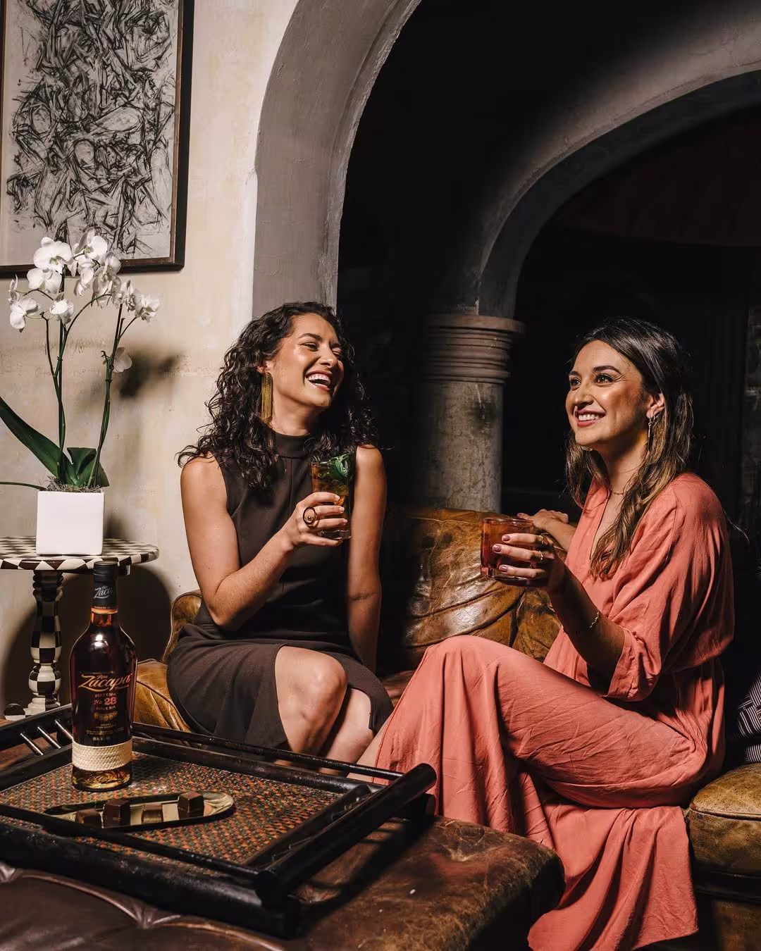 Two women sitting on a couch and enjoying cocktails, with a bottle of Mr Black on a side table and a white orchid in the background