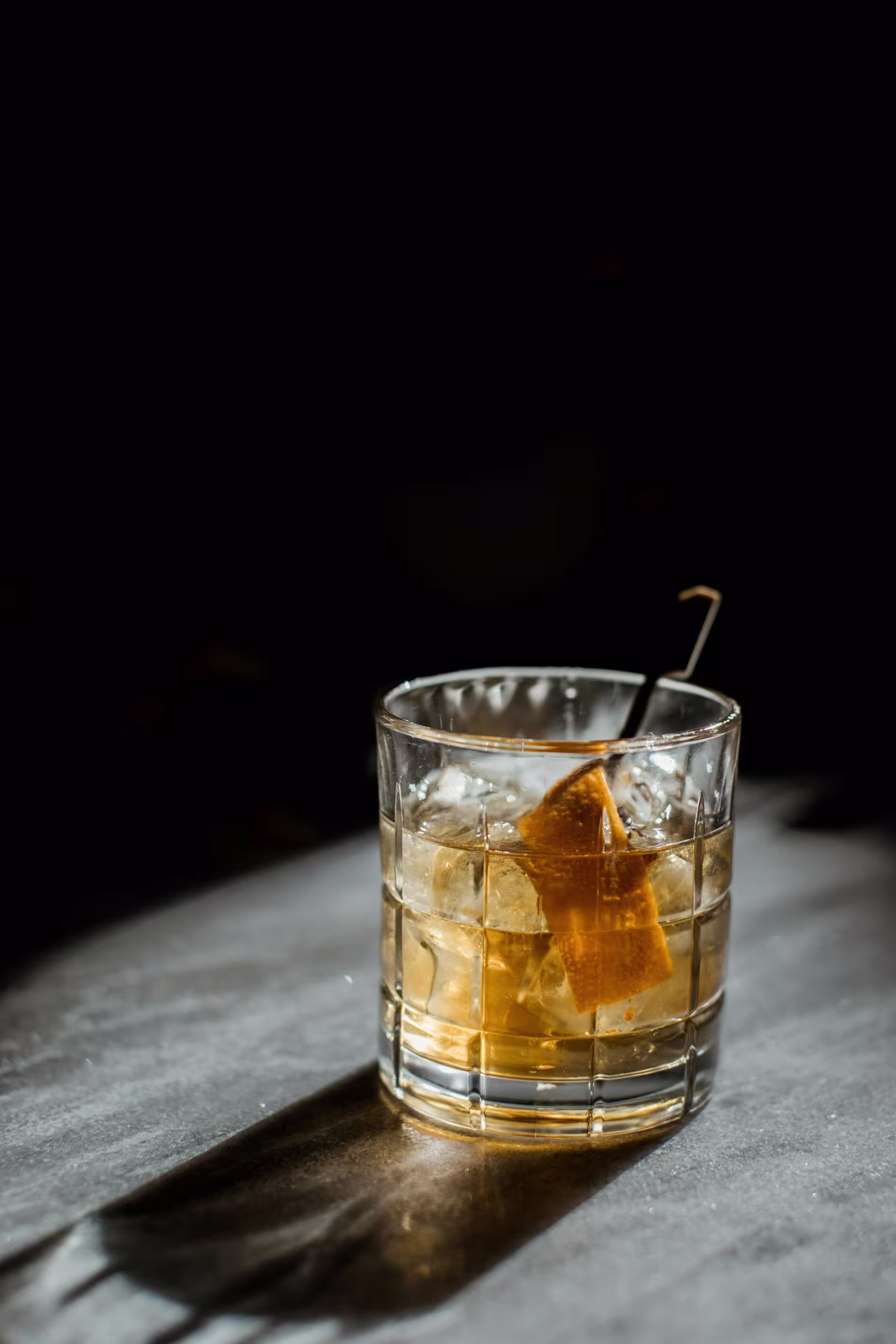 A glass of whiskey with ice cubes and an orange peel garnish sits on a sunlit gray surface against a dark background.