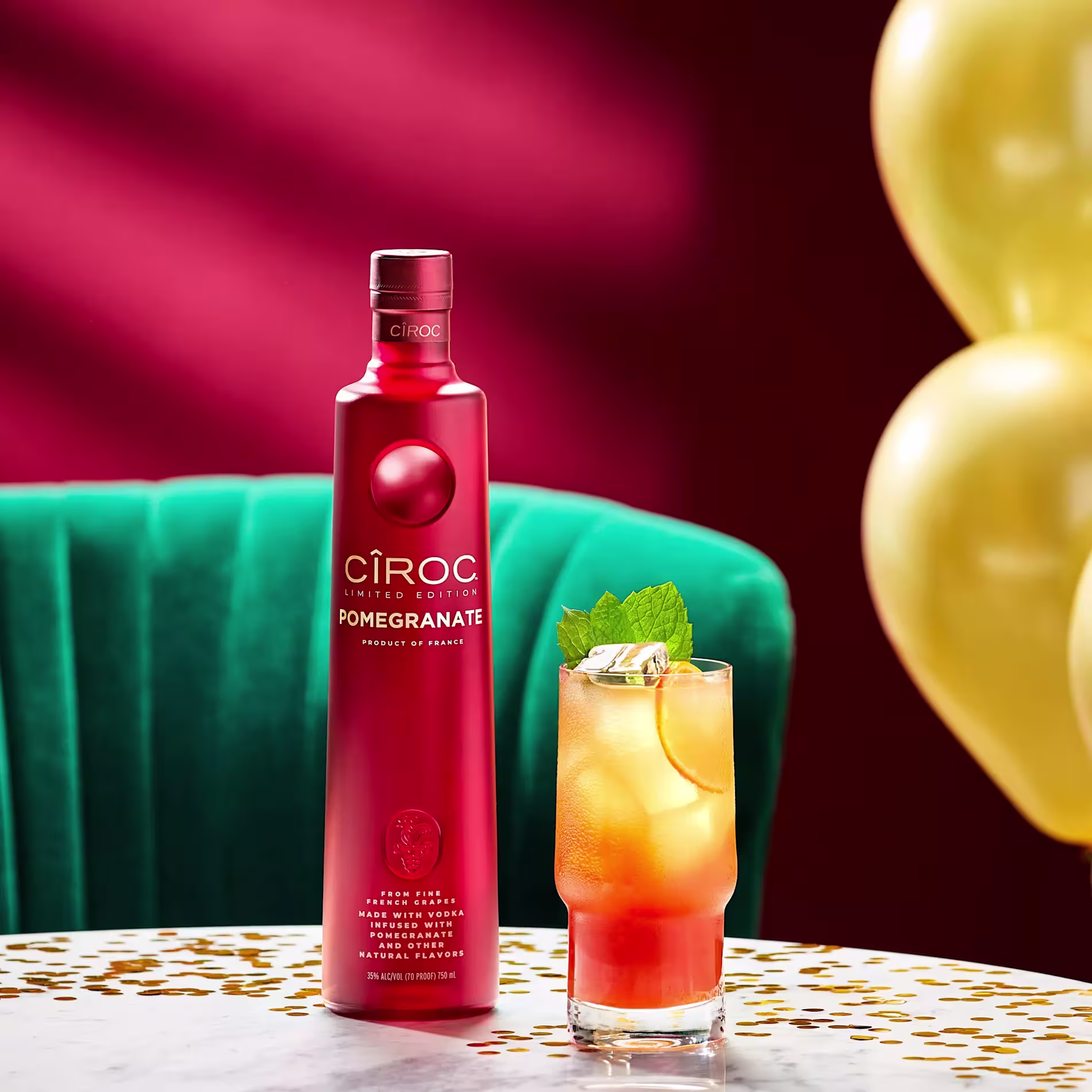 A red bottle of Cîroc Pomegranate vodka stands next to a cocktail with ice, citrus slices, and mint on a marble table, with a green chair and gold balloons in the background.