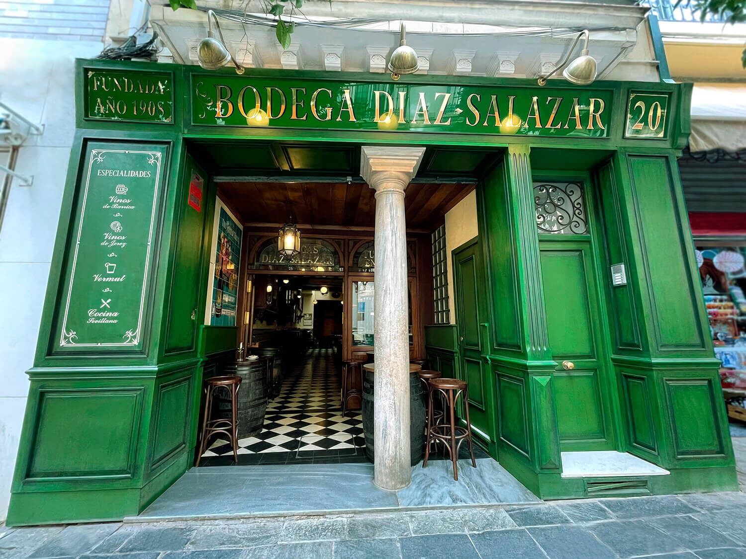 Fachada verde icónica de Bodega Díaz Salazar, con su pilar central, azulejos clásicos y barriles que invitan a entrar a una taberna con historia desde 1908.