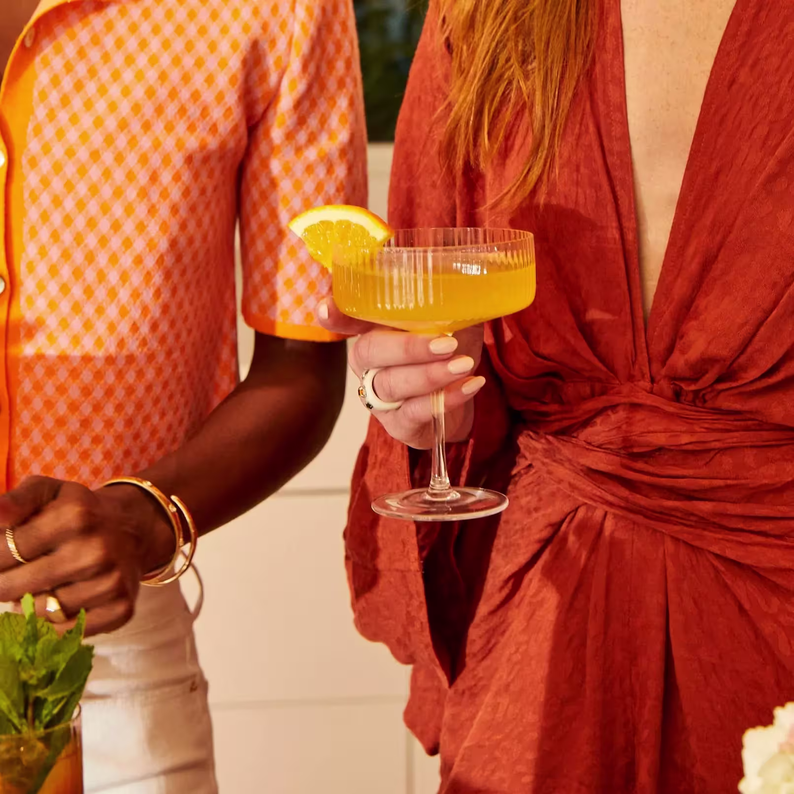 Two people in orange-toned outfits hold cocktails; one glass is garnished with an orange slice.