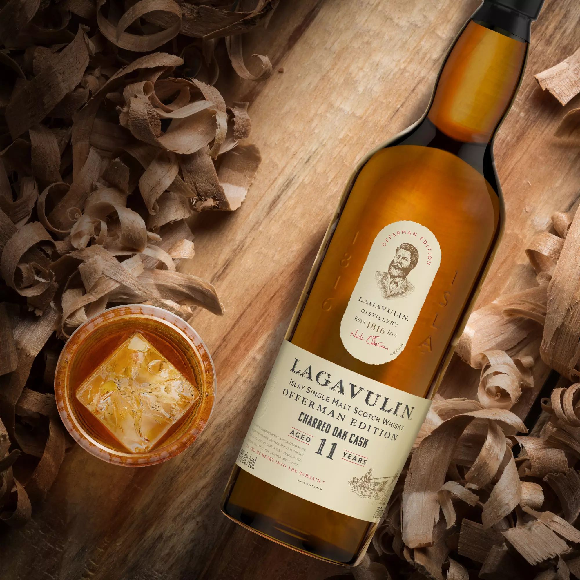A bottle of Lagavulin 11 Year Old Islay Single Malt Scotch Whisky lies on wood shavings next to a glass of whisky with ice.