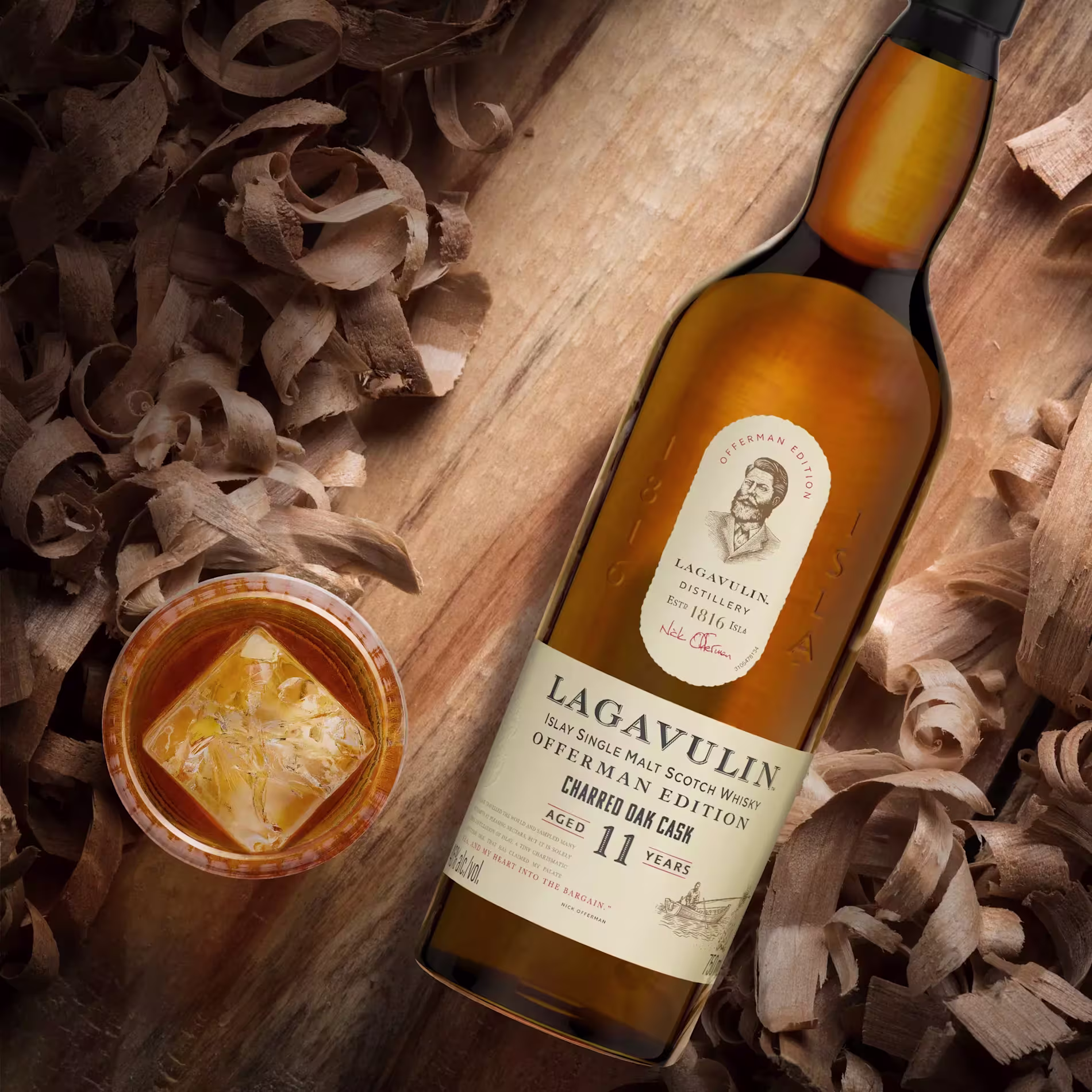A bottle of Lagavulin 11 Year Old Islay Single Malt Scotch Whisky lies on wood shavings next to a glass of whisky with ice.