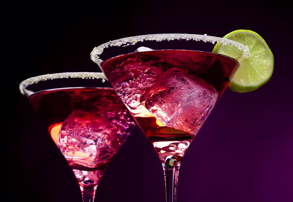 Two martini glasses filled with a red cocktail, each with ice cubes, a sugared rim, and a lime slice garnish, set against a dark purple background.