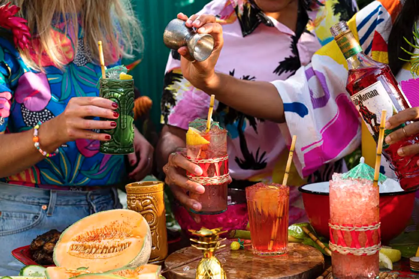 Three people prepare colorful tropical cocktails at an outdoor table decorated with fruit, drinks, and festive accessories.