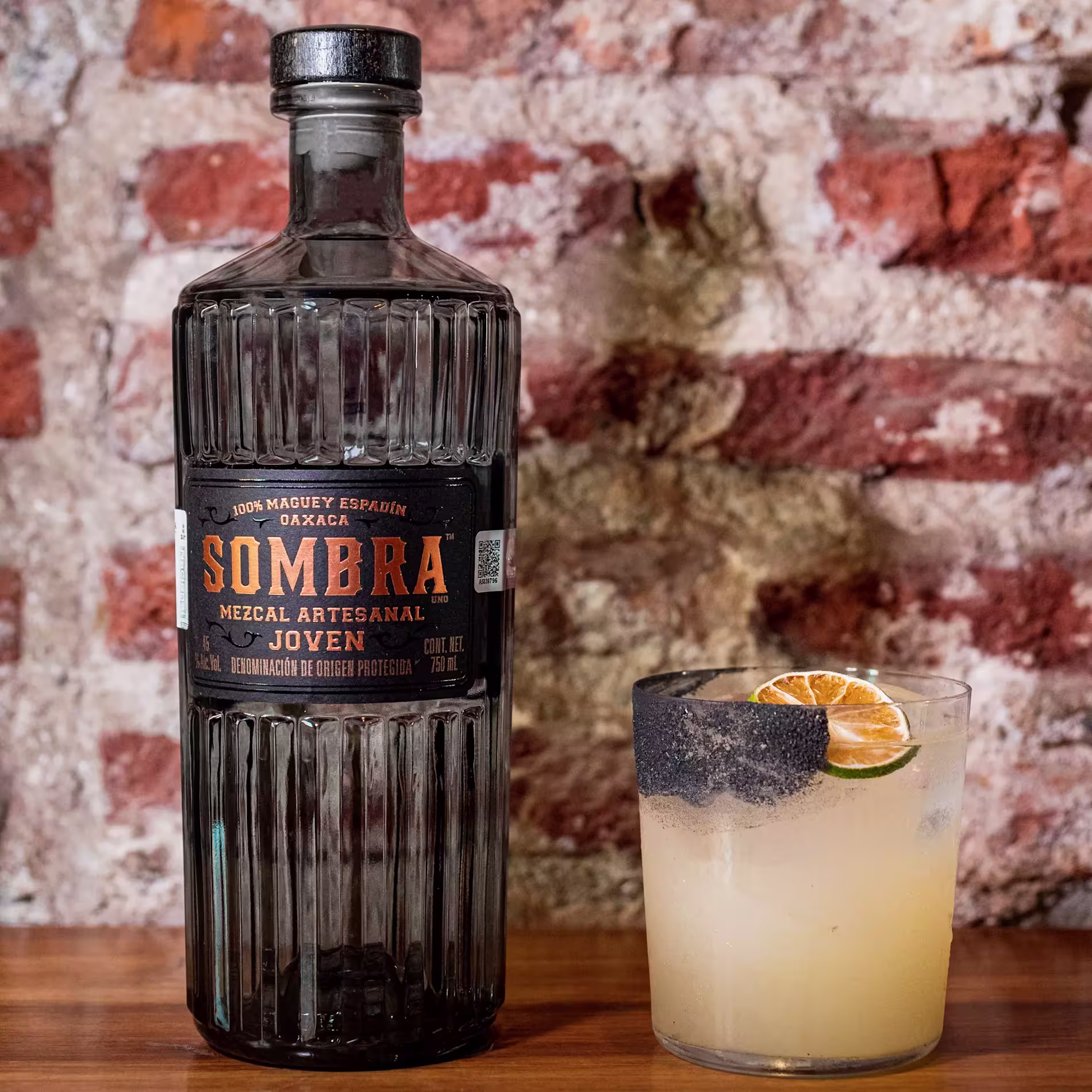 A bottle of Sombra Mezcal Artesanal stands next to a cocktail garnished with a lime wheel and black salt rim, set against a rustic brick wall.