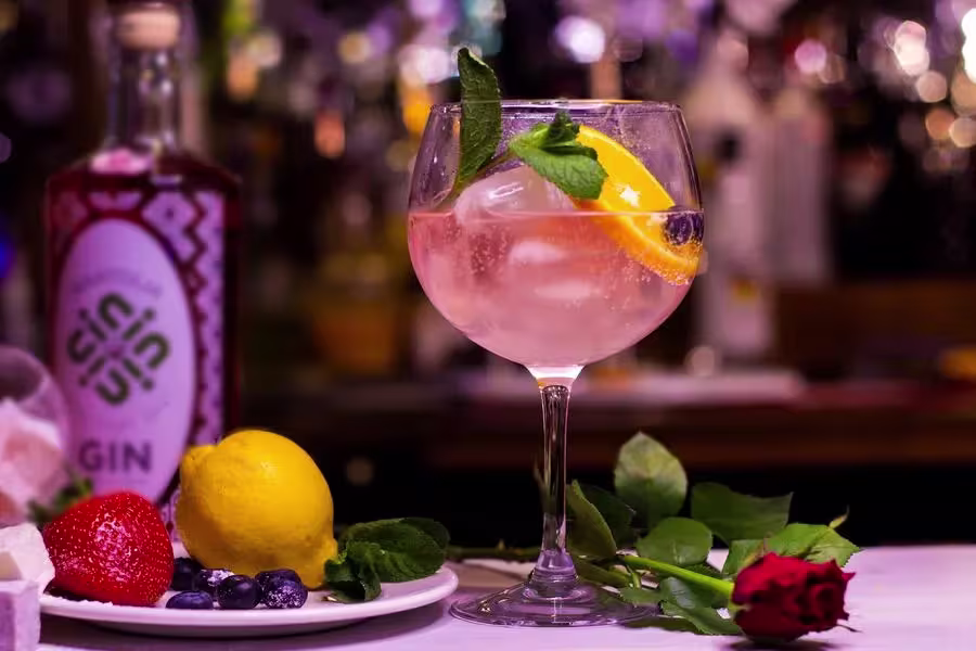 A glass of gin cocktail with ice, orange slice, mint, and a blueberry, beside a gin bottle, lemon, strawberry, blueberries, and a red rose on a bar counter.