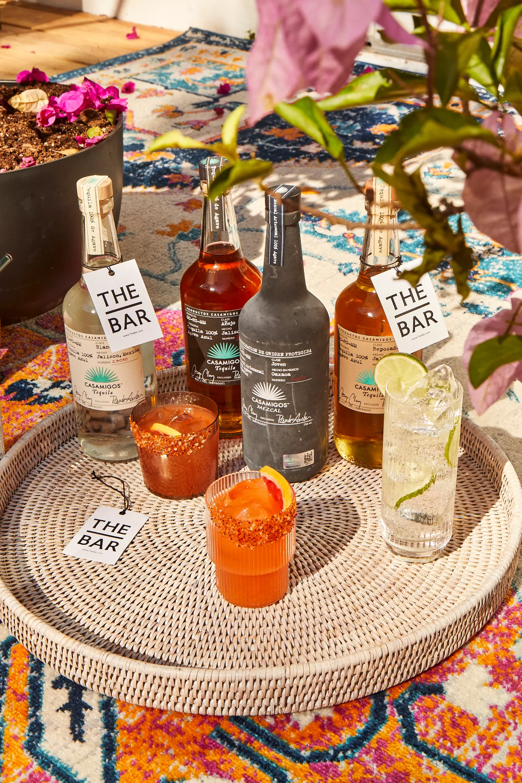 Four bottles of liquor, three mixed drinks, and two “The Bar” tags are arranged on a woven tray placed on a colorful patterned rug, with plants visible in the background.