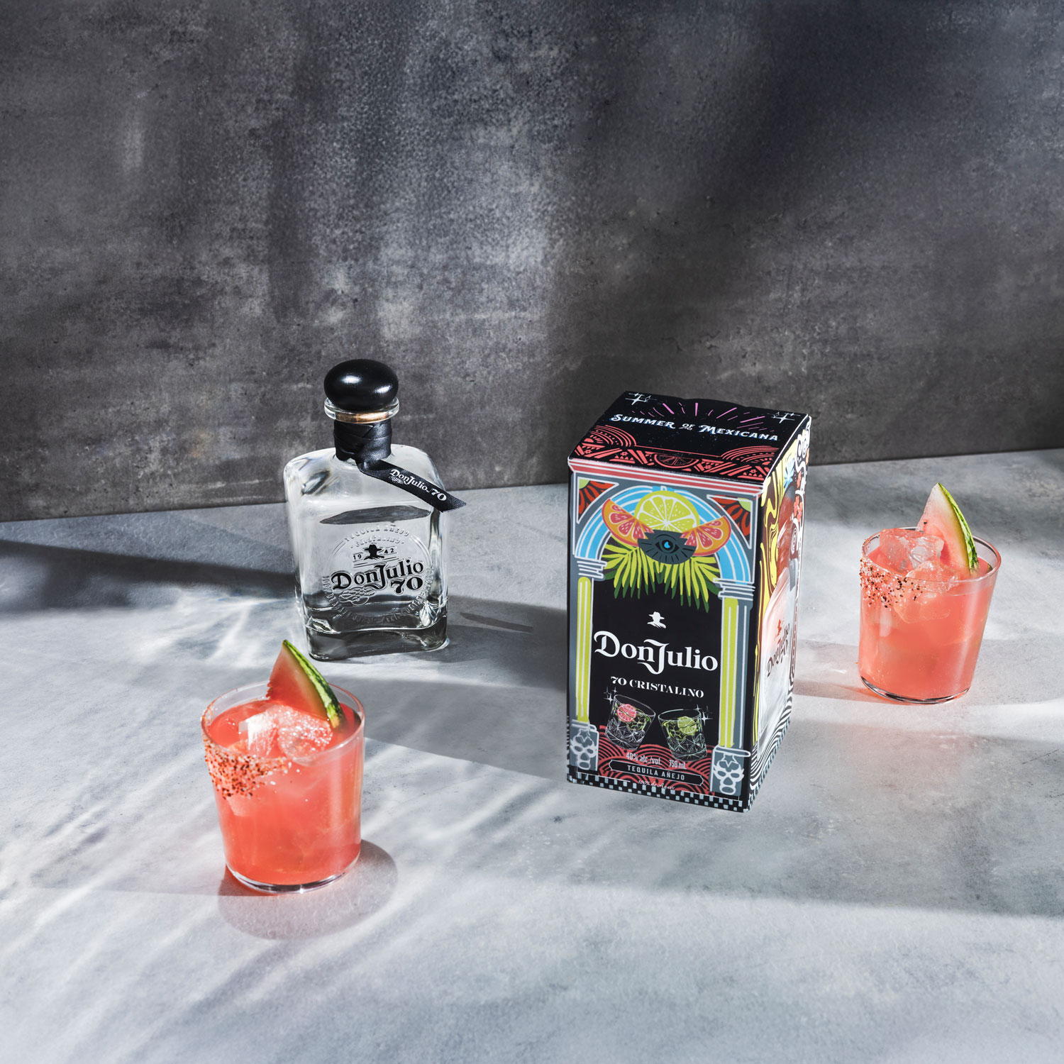 A bottle of Don Julio tequila, a decorative box, and two pink cocktails with ice, chili rims, and lime wedges on a gray surface.