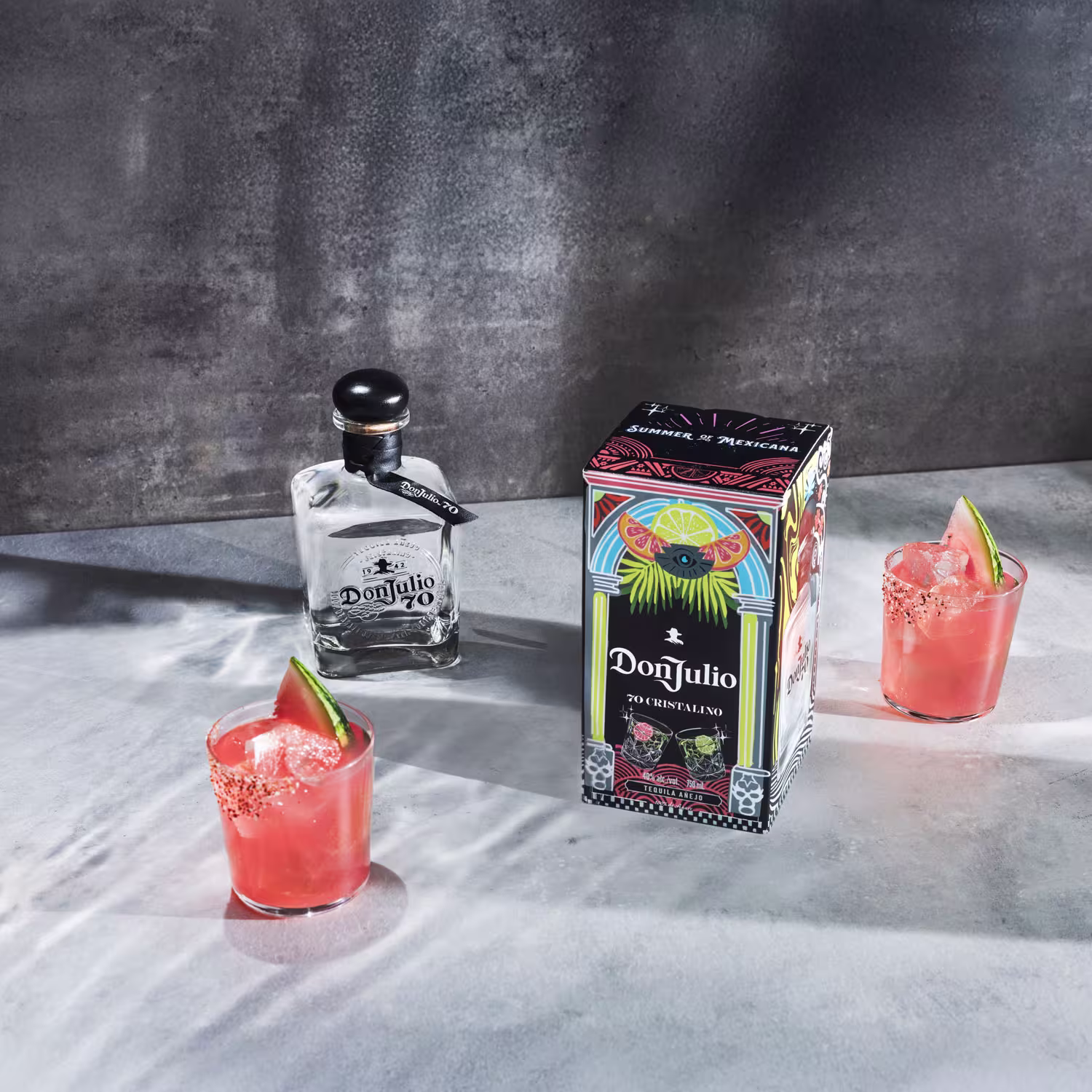 A bottle of Don Julio tequila, a decorative box, and two pink cocktails with ice, chili rims, and lime wedges on a gray surface.