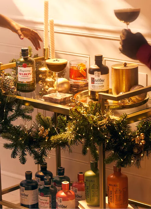 A decorated bar cart with various bottles, cocktails, and holiday greenery; two people reach for drinks, one holding a coupe glass.