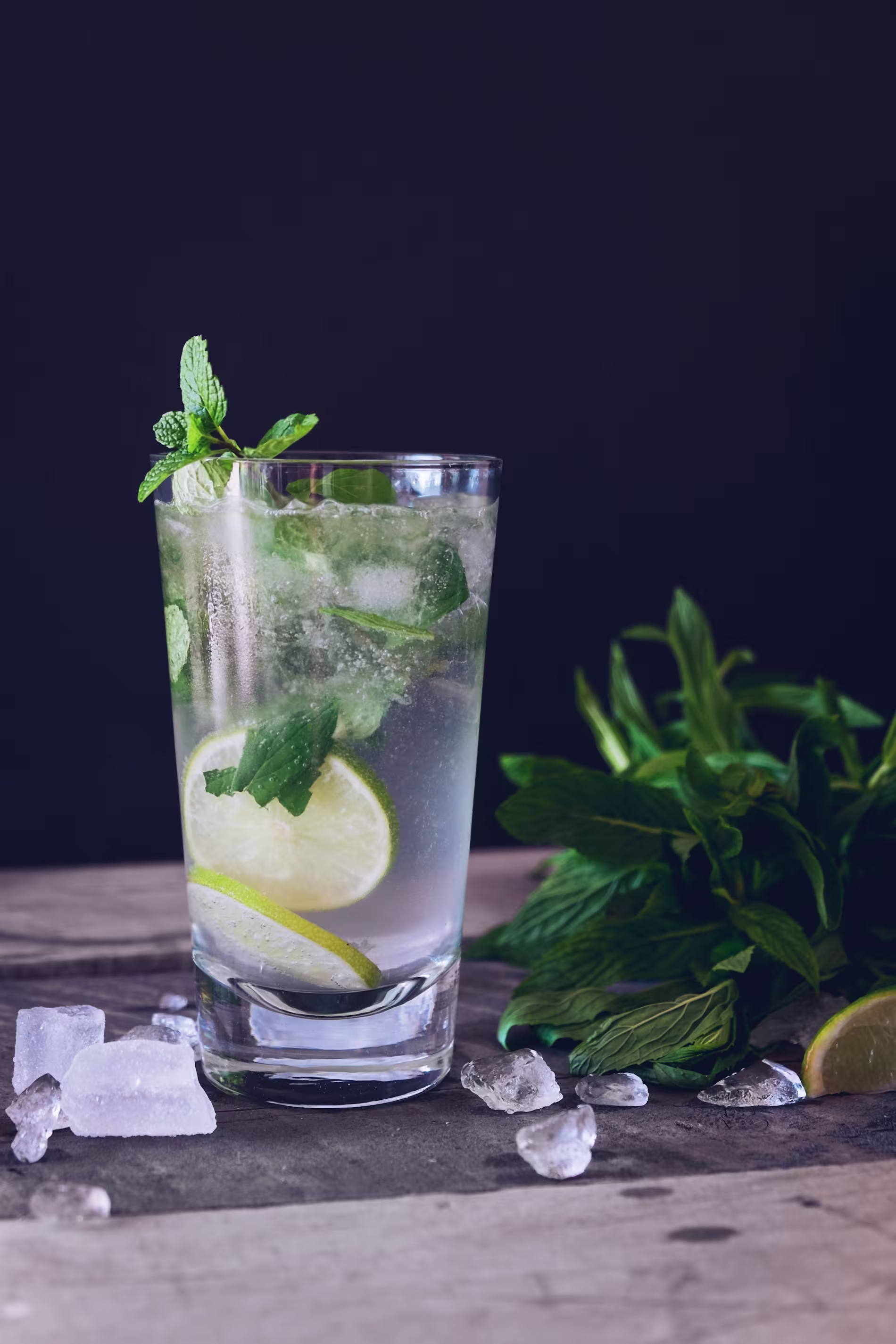 A glass of clear drink with lime slices and mint, surrounded by ice cubes and fresh mint leaves on a wooden surface.