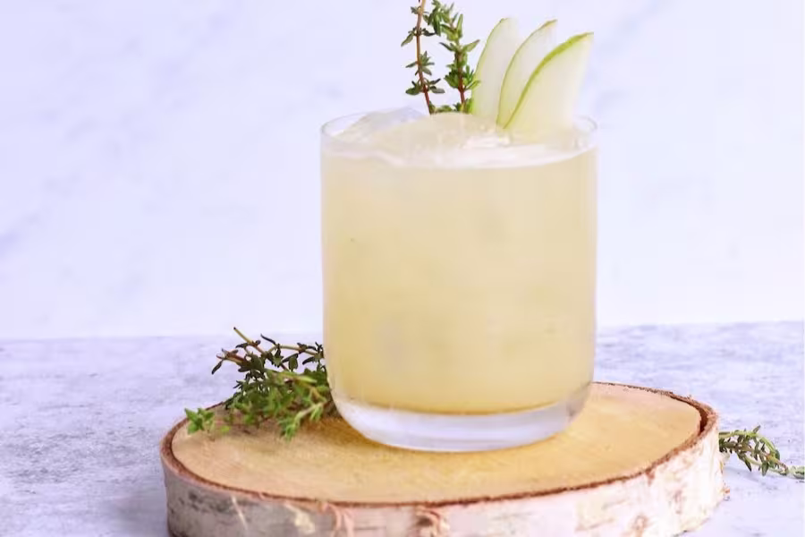 A glass of light yellow cocktail with ice, garnished with pear slices and a sprig of thyme, sits on a wooden coaster against a light background.