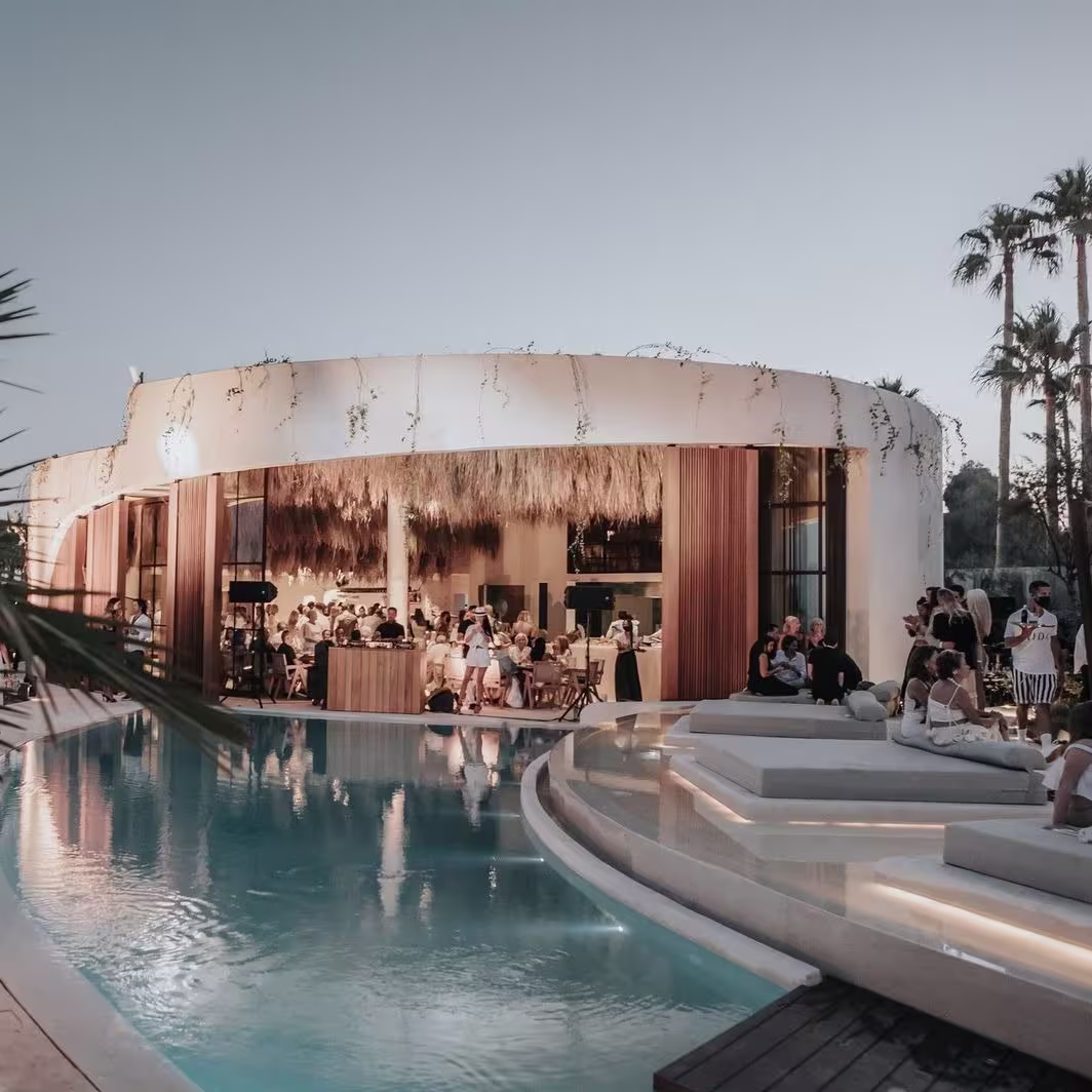 Exterior de Nido Beach Club en Estepona, con una piscina cristalina rodeada de zonas de descanso y una estructura moderna con techo de paja.