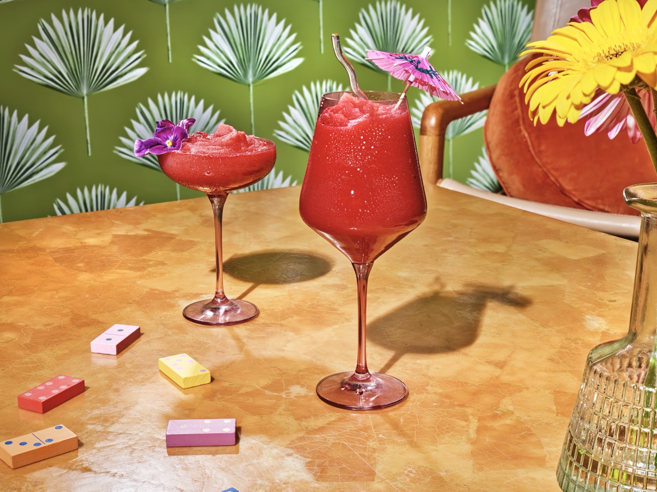 Two red frozen cocktails in stemmed glasses with garnishes sit on a tan table beside dominoes and a vase of yellow and orange flowers, with a green palm-patterned background.