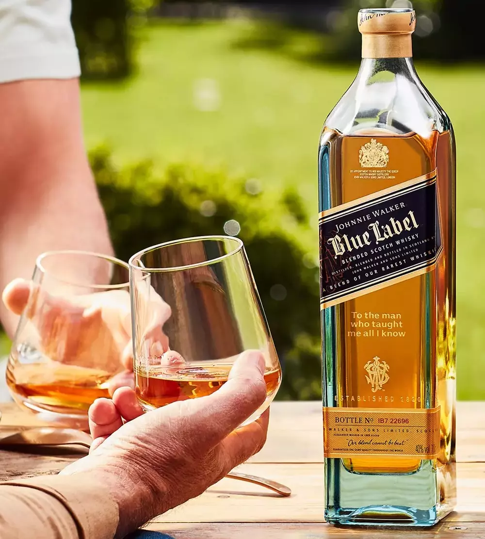 A bottle of Johnnie Walker Blue Label whisky is on a table outdoors, with two people holding glasses of whisky in the background.