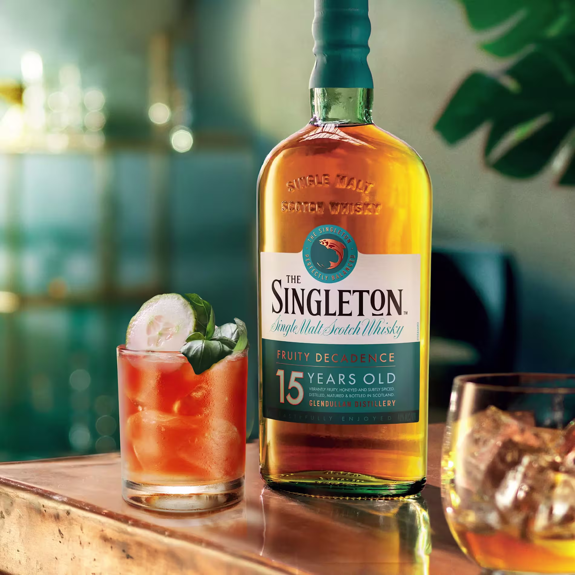 A bottle of The Singleton 15 Years Old single malt scotch whisky on a table, next to a cocktail with ice and garnish, and another glass with whisky and ice.