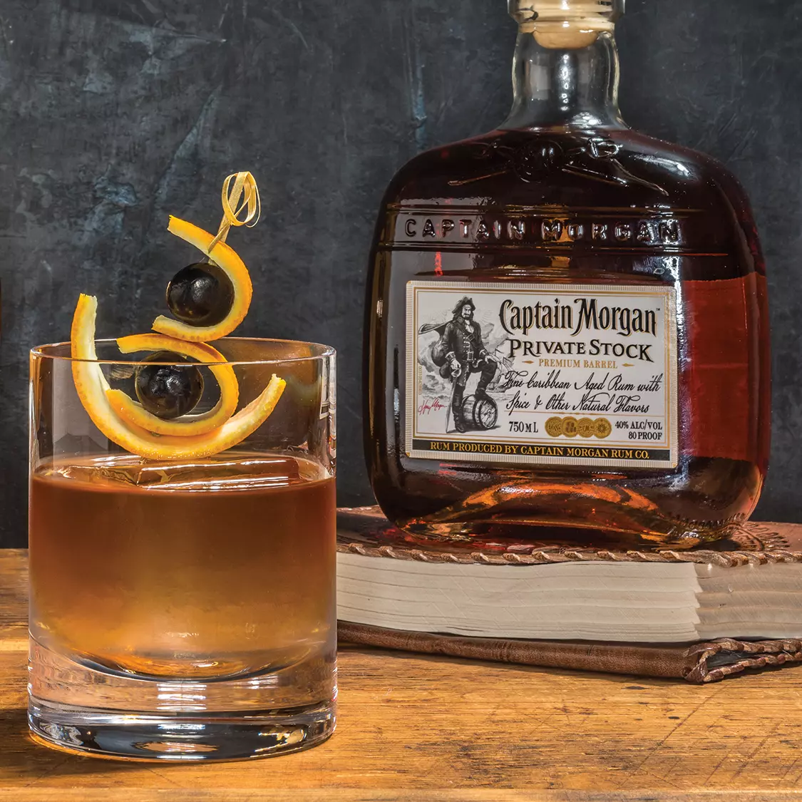 A glass of cocktail garnished with orange peel and cherries sits on a table next to a bottle of Captain Morgan Private Stock rum and a closed book.