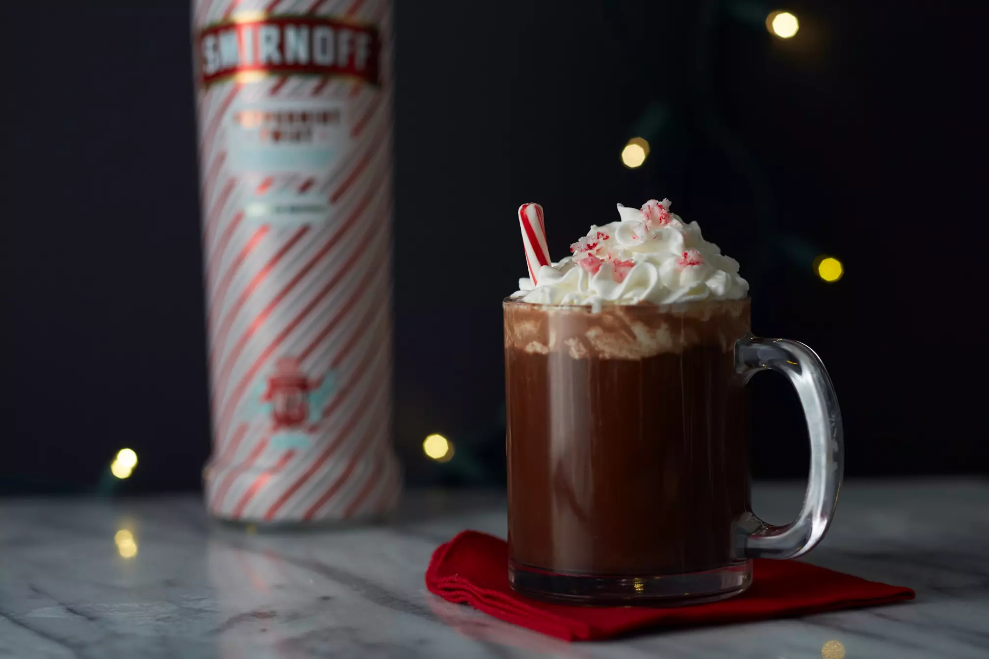 A glass mug of hot chocolate topped with whipped cream and crushed peppermint, garnished with a mini candy cane, sits in front of a bottle of Smirnoff Peppermint Twist.