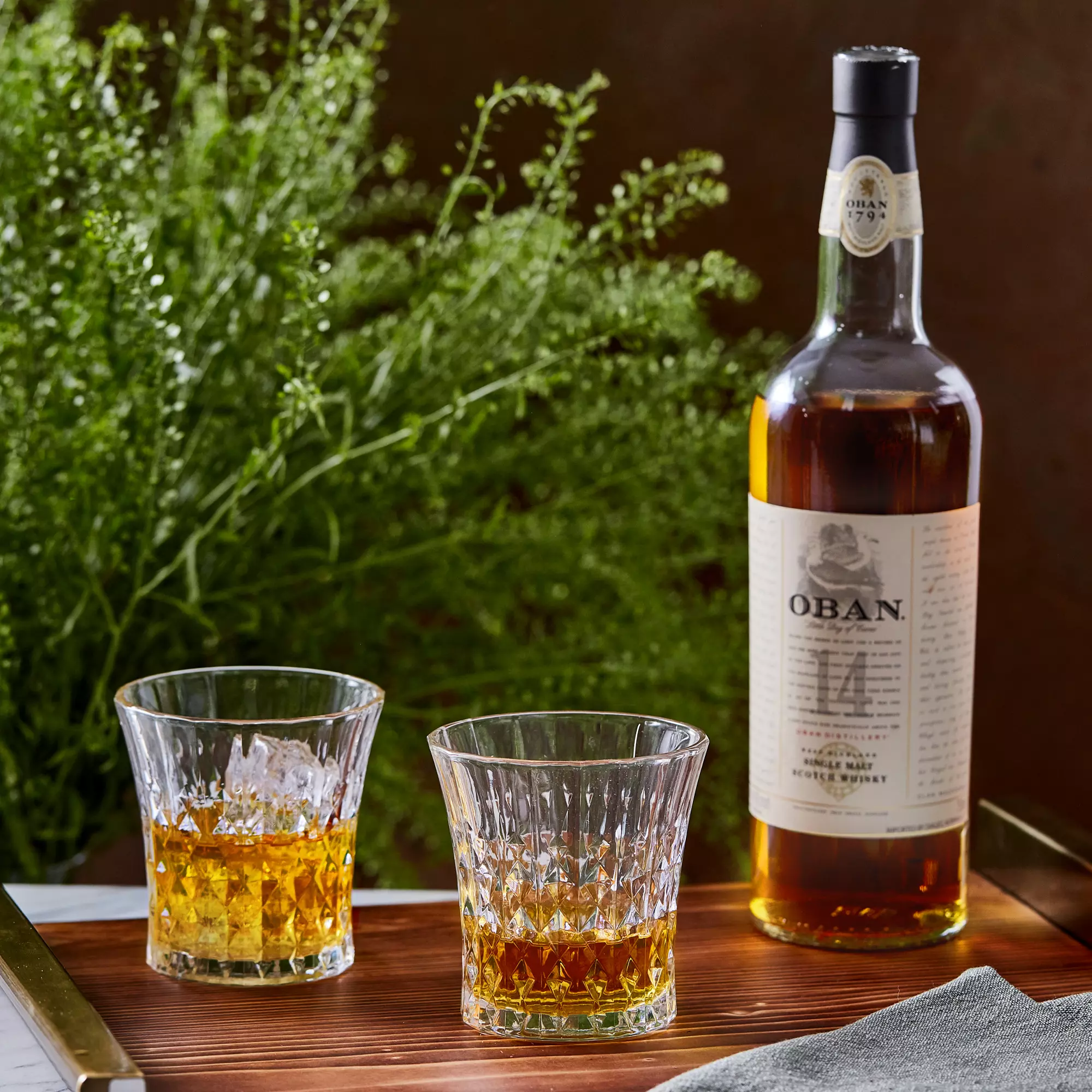 A bottle of Oban 14 Year Old Scotch whisky stands next to two glasses filled with whisky on a wooden tray, with green foliage in the background.