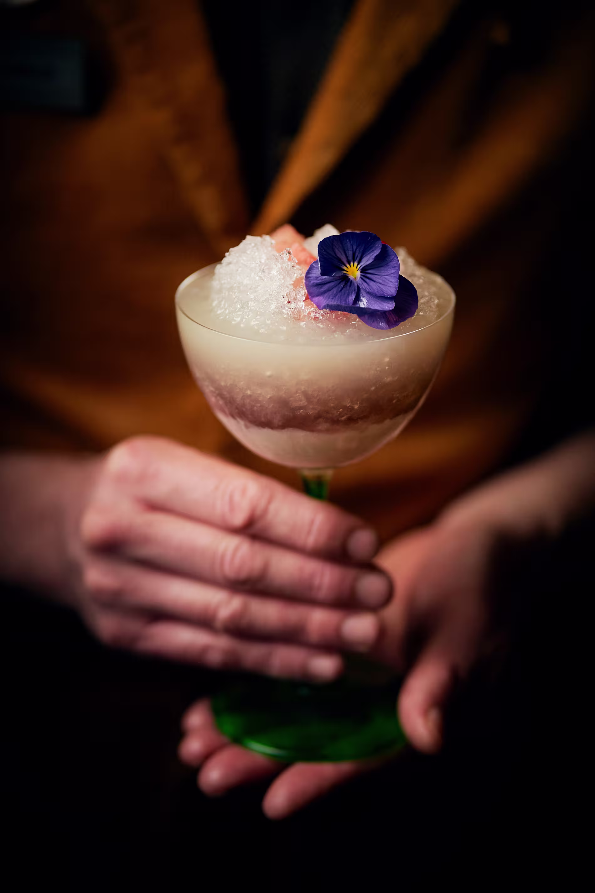 La imagen muestra un primer plano de una copa de cóctel con tallo, que está sobre una barra con un surtido de botellas desenfocado en el fondo. El cóctel tiene una capa espumosa en la parte superior y está adornado con hielo triturado y una flor morada.