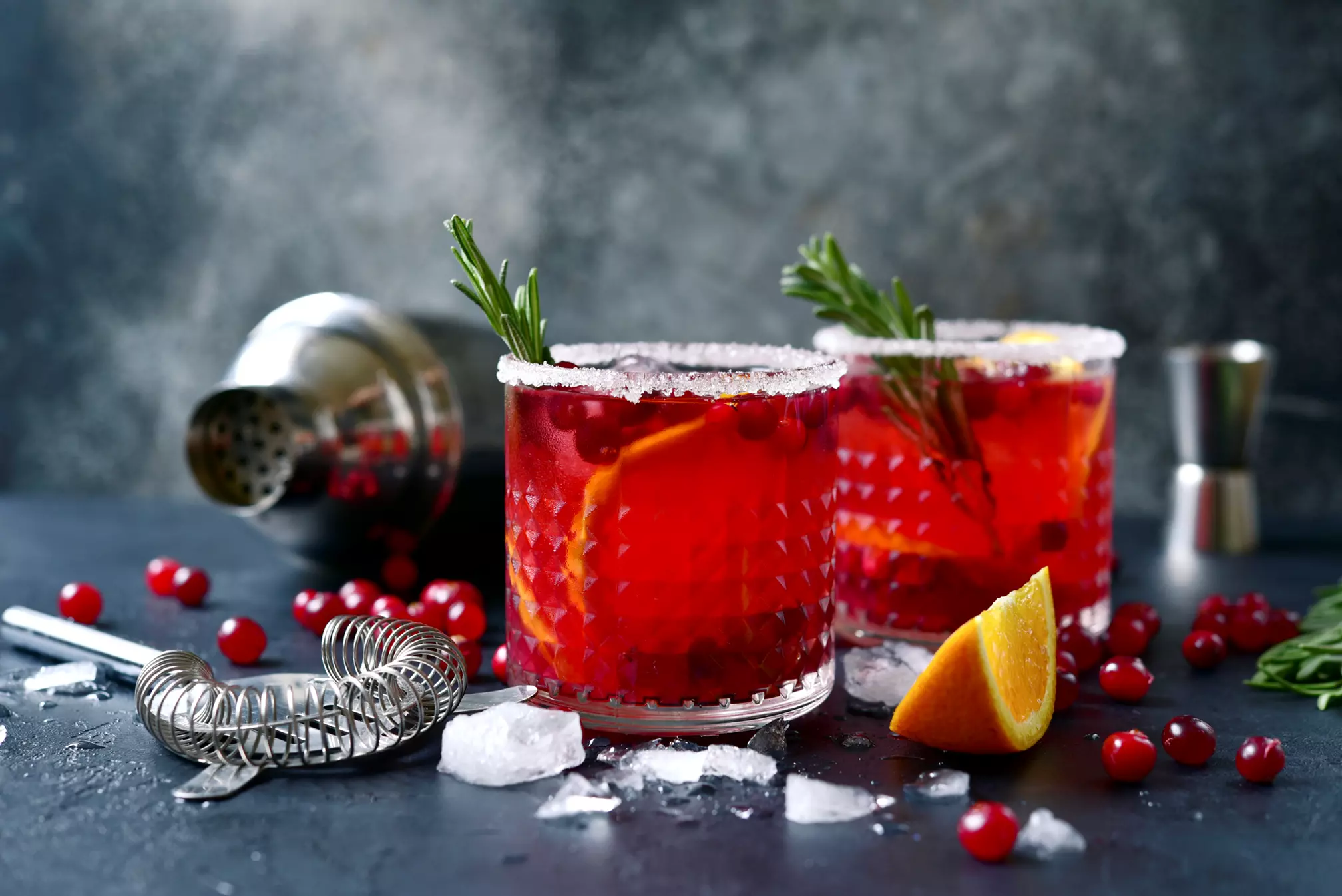 Two glasses of red cocktail garnished with orange slices and rosemary, surrounded by ice, cranberries, a cocktail strainer, shaker, and jigger on a dark surface.