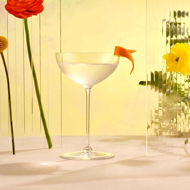 Aviation Gin martini in a coupe glass garnished with an orange twist, surrounded by colorful flowers on a light background