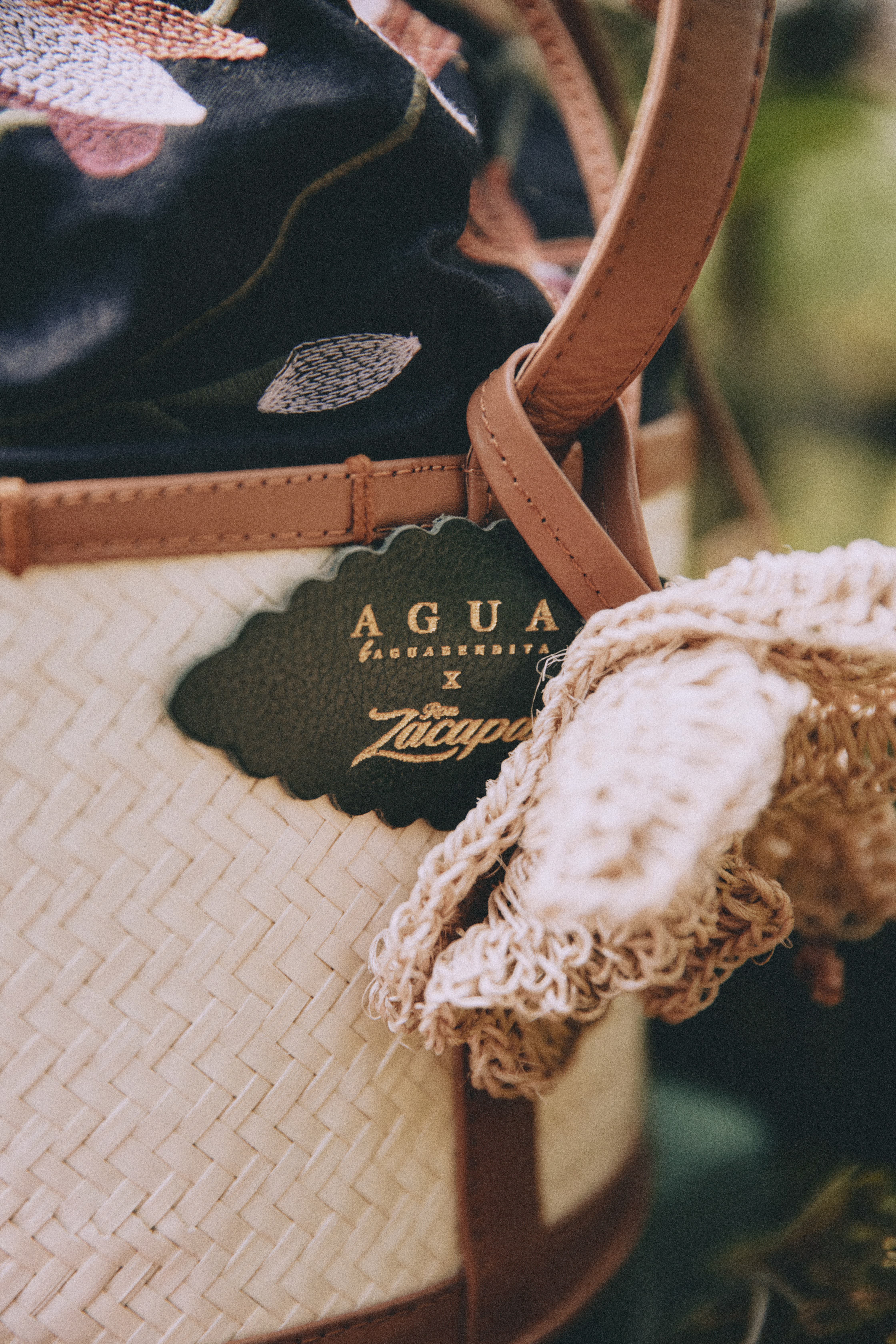 Close-up of a woven basket bag with brown leather accents, featuring a black and gold tag labeled AGUA x Zacapa and a decorative fabric flower attached.