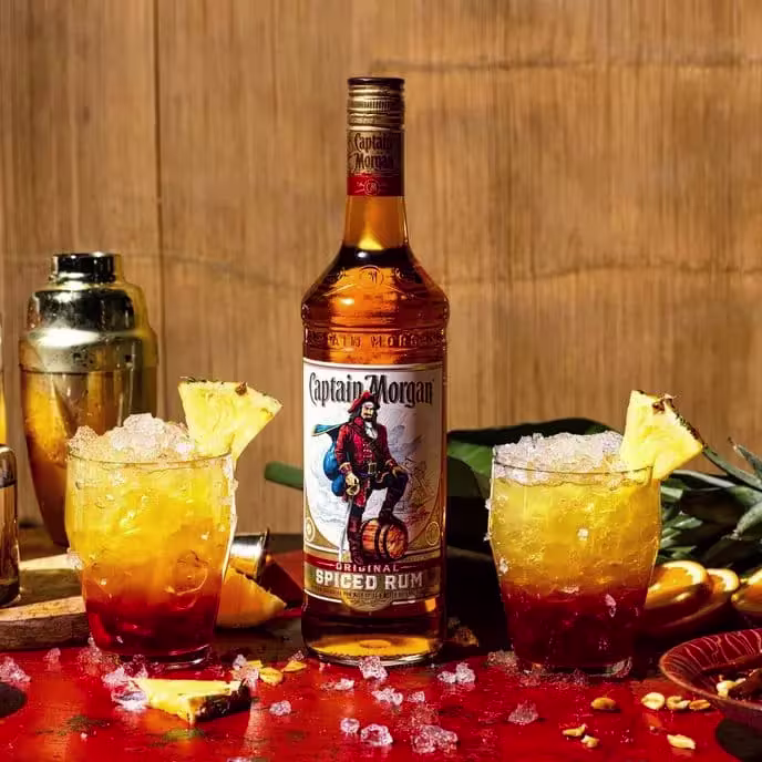 A bottle of Captain Morgan Original Spiced Rum is placed between two rum cocktails with pineapple garnishes on a red table, with a shaker and sliced fruit nearby.