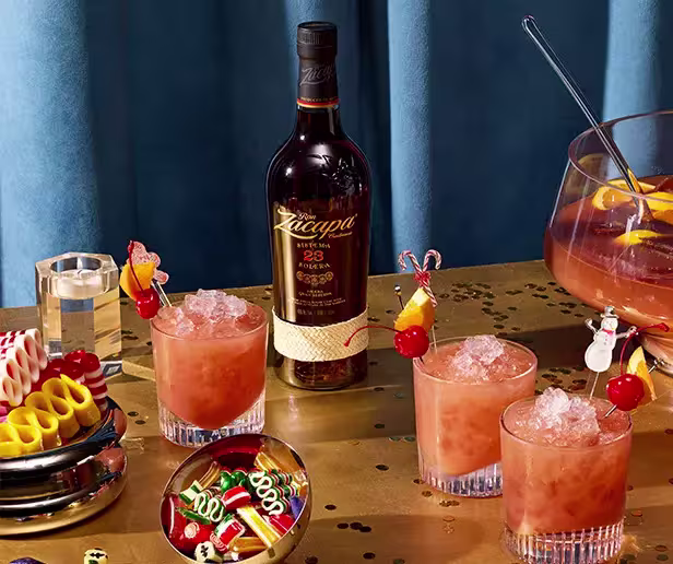 A bottle of Zacapa rum and three glasses of orange-pink cocktails with ice are arranged on a table with party snacks, confetti, and a punch bowl.