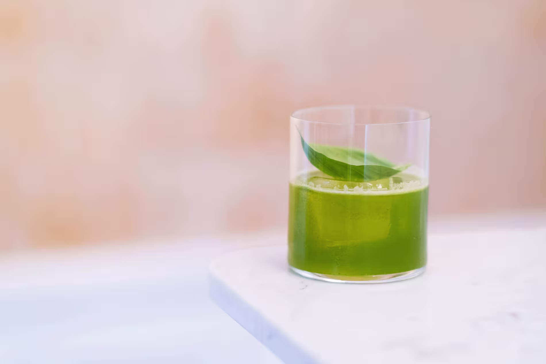 A clear glass filled with green juice, garnished with a single leaf, sits on a white marble surface against a soft beige background.