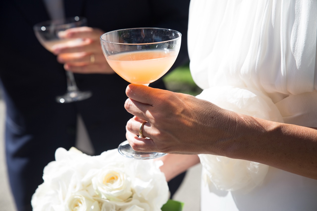 A person in a white outfit holds a cocktail glass and a bouquet of white roses, with another person holding a similar drink in the background.