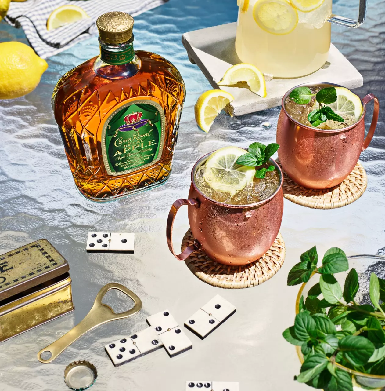 A bottle of Crown Royal apple whiskey, two copper mugs with drinks and lemon garnish, a pitcher of lemonade, dominoes, a bottle opener, and fresh mint on a glass table.