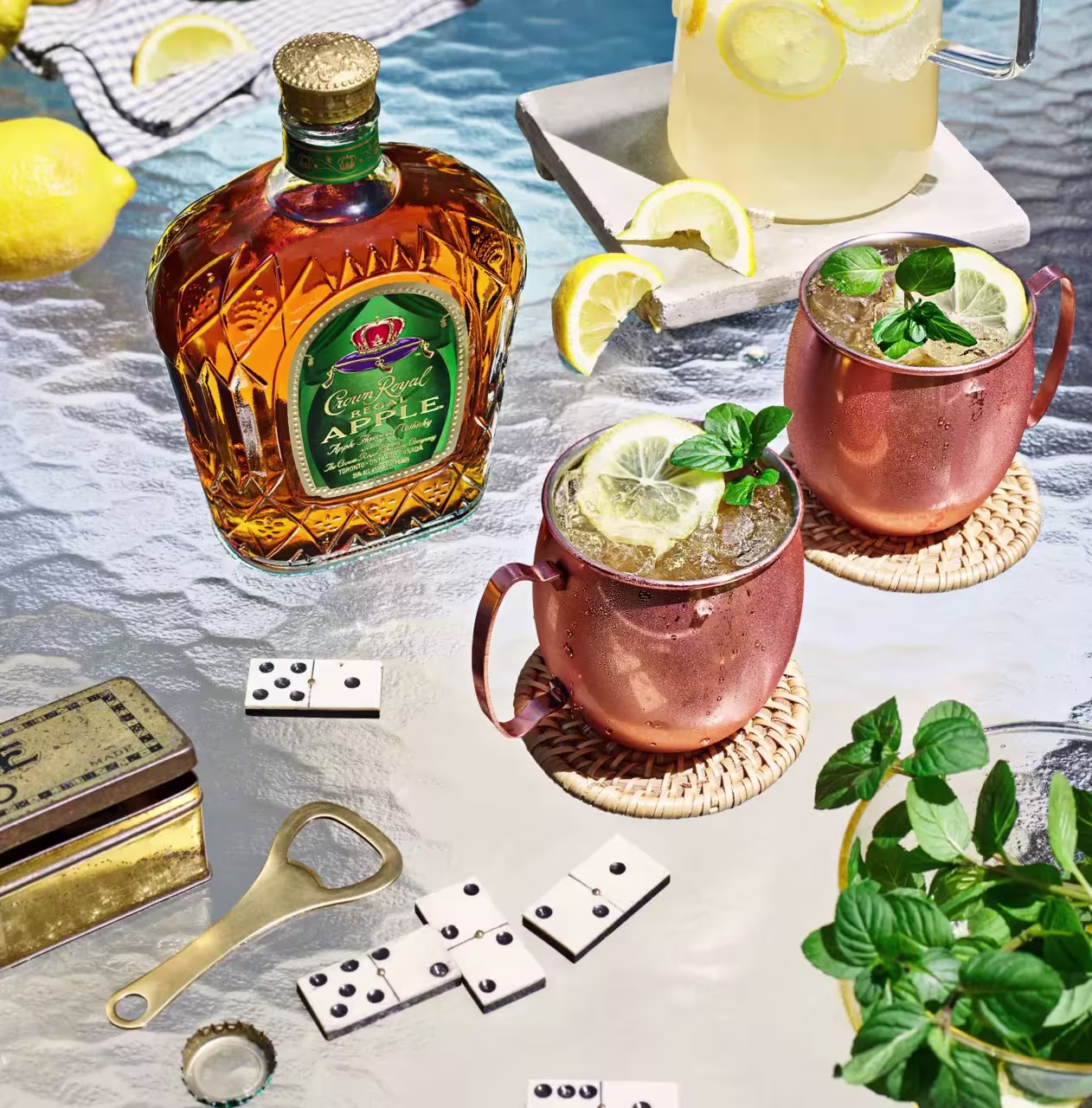 A bottle of Crown Royal apple whiskey, two copper mugs with drinks and lemon garnish, a pitcher of lemonade, dominoes, a bottle opener, and fresh mint on a glass table.