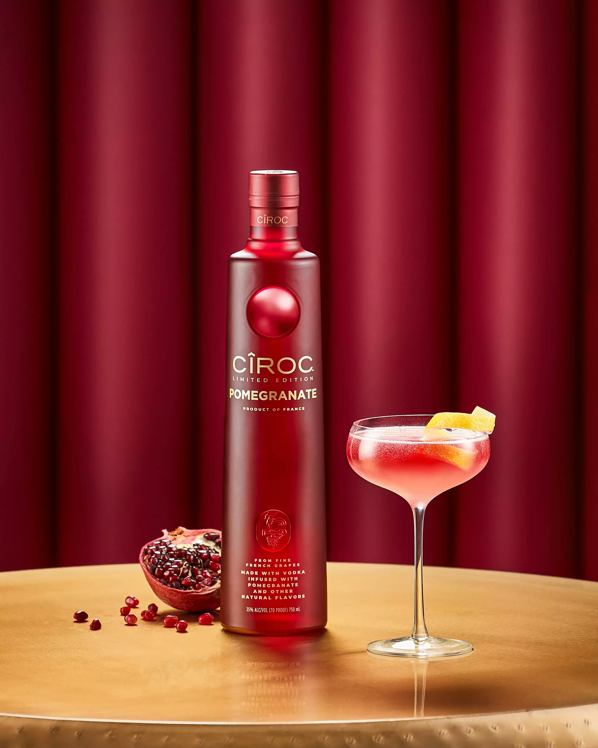 A bottle of Cîroc Pomegranate vodka stands on a gold table next to a cocktail glass with a lemon garnish and a halved pomegranate with seeds, against a red background.
